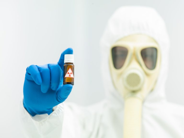 20691577 - radioactive isotope sample in a small brown bottle with a warning label held in the fingers of a laboratory scientist in full protective clothing with a mask and breathing apparatus