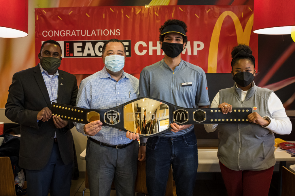 Four people hold up the McDonald's Crew League winning belt in a McDonald's restaurant